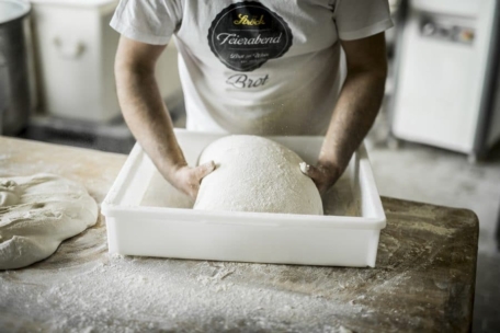 Eine Person in einem weißen T-Shirt knetet eine große Teigkugel in einem weißen Plastikbehälter auf einer bemehlten Holzfläche in einer Bäckerei.