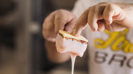 Eine Person hält einen Keks, der teilweise in dicke, weiße Glasur getaucht ist, wobei die Glasur vom Rand herabtropft. Der Hintergrund ist unscharf und konzentriert sich auf den Keks und die Hände der Person.
