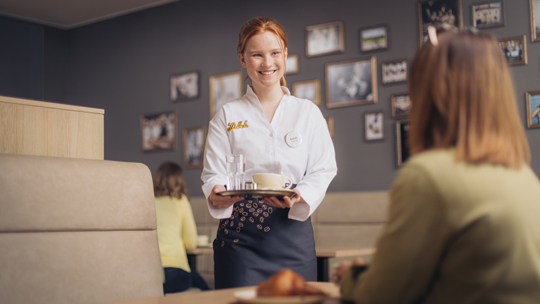 Eine lächelnde Kellnerin in Uniform serviert einem sitzenden Kunden in einem gemütlichen Restaurant mit gerahmten Bildern an der Wand ein Getränk und eine Schale auf einem Tablett.