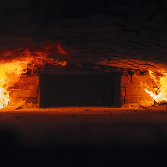 In einem gemauerten Ofen brennen auf jeder Seite helle Flammen, die das dunkle Innere in ein intensives orangefarbenes Licht tauchen. Die gewölbte Decke und das Mauerwerk sind in der Hitze sichtbar.