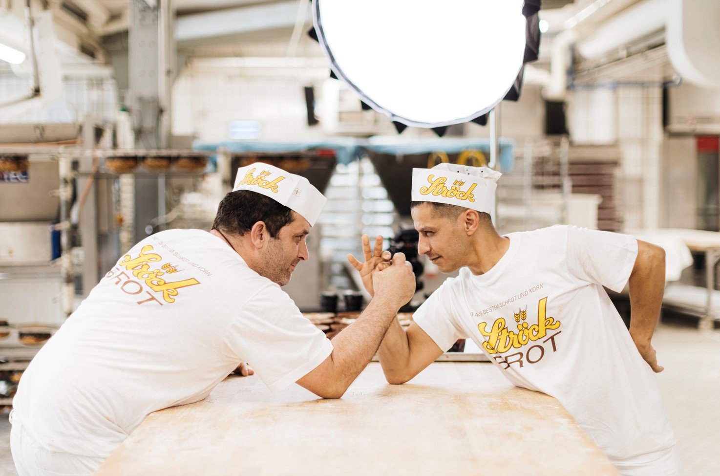 Zwei Bäcker in weißen Uniformen und Hüten ringen auf einem Holztisch in einer Bäckerei mit den Armen, lächeln sich an und schauen sich an, während im Hintergrund Backgeräte und helle Beleuchtung zu sehen sind.