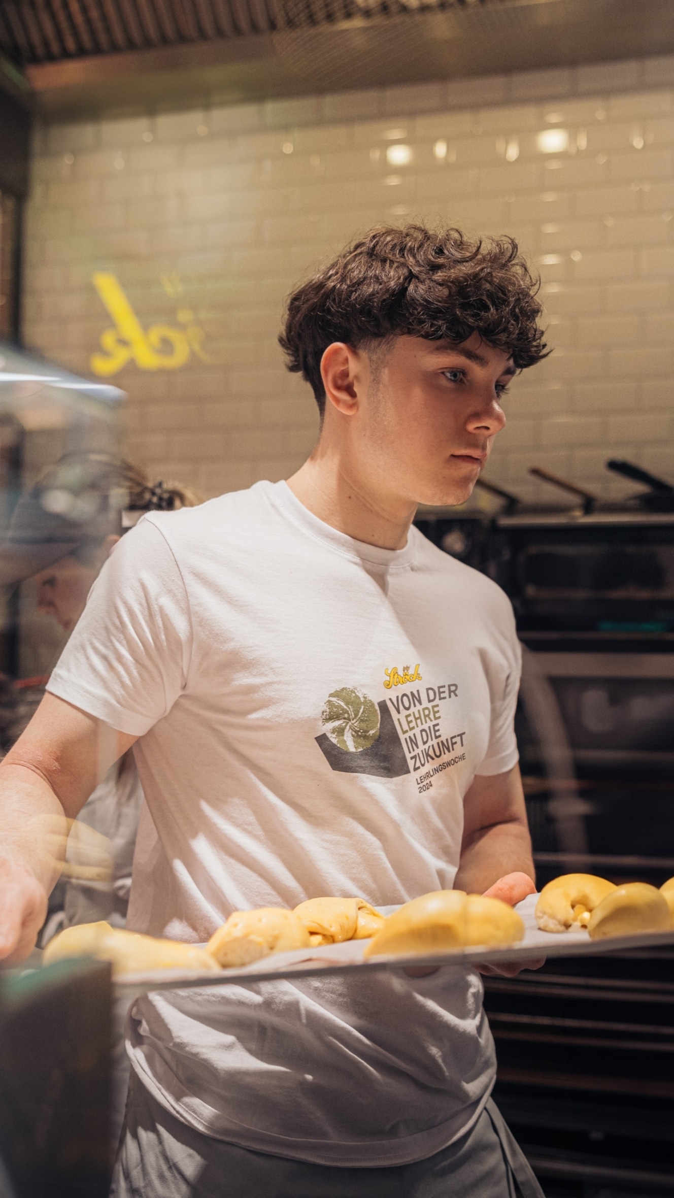 Ein junger Mann mit lockigem Haar, der ein weißes T-Shirt mit deutscher Aufschrift trägt, steht hinter einem Tresen mit einem Tablett voller Gebäck in einer Bäckerei oder einem Café mit weiß gekachelten Wänden und Küchengeräten im Hintergrund.