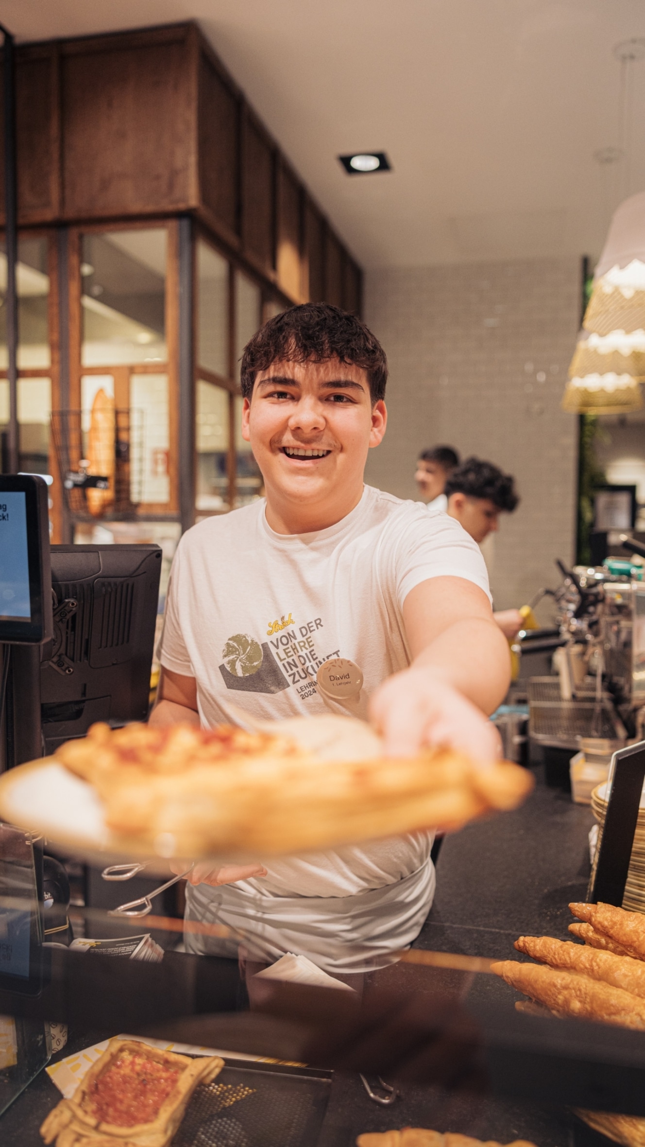Eine lächelnde junge Person hinter einer Bäckertheke reicht einen Teller mit Gebäck in Richtung der Kamera. Auf der Theke sind Gebäck und Backwaren zu sehen, im Hintergrund sind weitere Mitarbeiter und moderne Dekoration zu sehen.