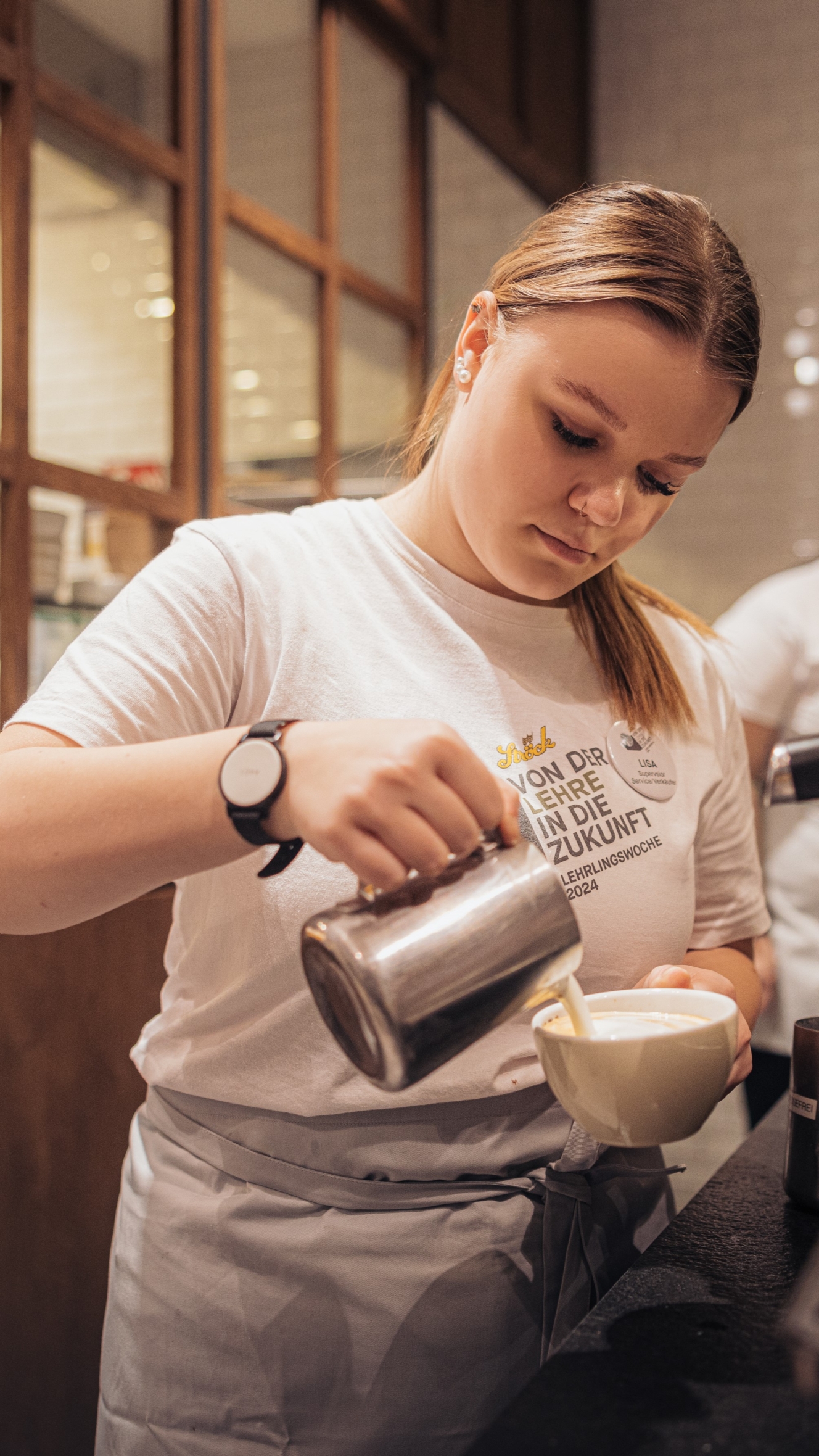 Eine junge Frau in einem weißen T-Shirt und einer Schürze gießt vorsichtig Milch aus einem Metallkrug in eine Tasse und bereitet ein Kaffeegetränk an einem Café-Tresen zu.